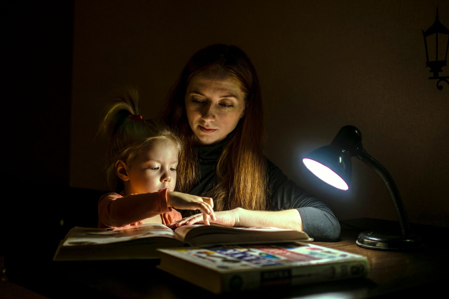A mother and daughter read a book together under a warm lamp light.